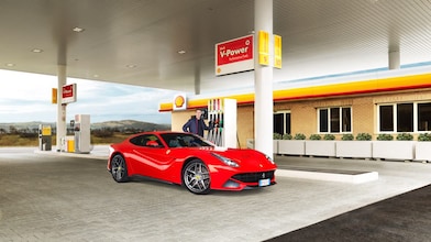 Une Ferrari rouge arrêtée à la pompe d'une station Shell avec un homme qui s'apprête à faire le plein