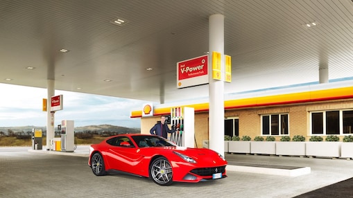 Une Ferrari rouge arrêtée à la pompe d'une station Shell avec un homme qui s'apprête à faire le plein