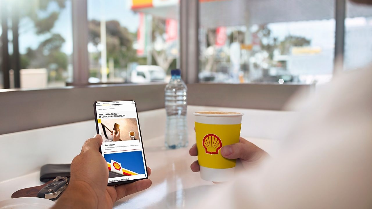 Client en train de consulter l'application mobile Shell en buvant un café dans une station Shell